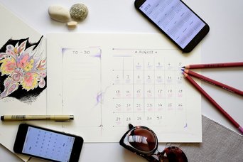 Auf einem Tisch liegen ein Kalender, Stifte, zwei Handys und eine Sonnenbrille